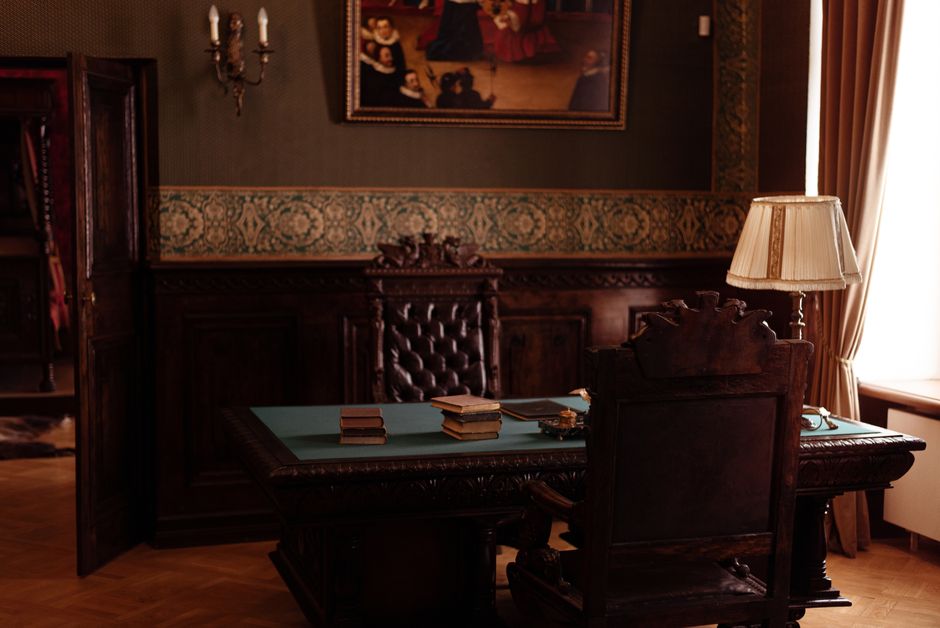 An ornate study room featuring antique wooden furniture, books, and a classic lamp.